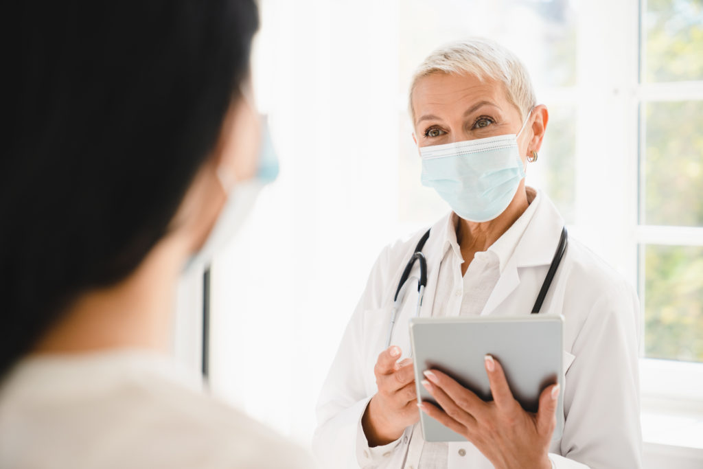 Doctor in protective face mask against Covid 19 using tablet talking with patient about diagnosis