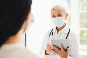 Doctor in protective face mask against Covid 19 using tablet talking with patient about diagnosis